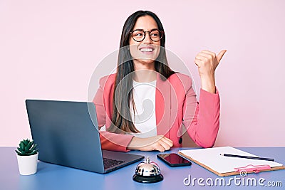 Young caucasian woman sitting at the recepcionist desk working using laptop smiling with happy face looking and pointing to the Stock Photo