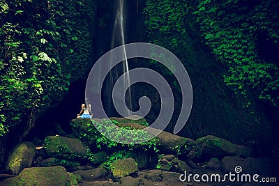 Young woman sitting on the rock, practicing yoga near waterfall. Hands in gyan mudra. Leke Leke waterfall, Bali. View from back Stock Photo