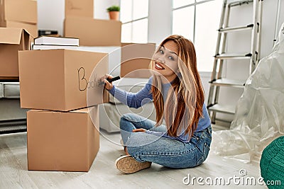 Young caucasian woman smiling confident writing books word on cardboard box at new home Stock Photo