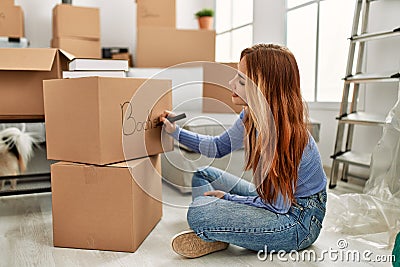 Young caucasian woman smiling confident writing books word on cardboard box at new home Stock Photo