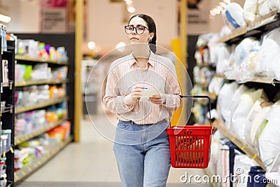 Young Caucasian woman wearing glasses holds grocery basket, holds list and selects bed linen and pillow. Shopping and Stock Photo