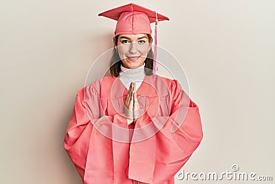 Young caucasian woman wearing graduation cap and ceremony robe praying with hands together asking for forgiveness smiling Stock Photo