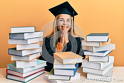 Young caucasian woman wearing graduation ceremony robe sitting on the table praying with hands together asking for forgiveness Stock Photo