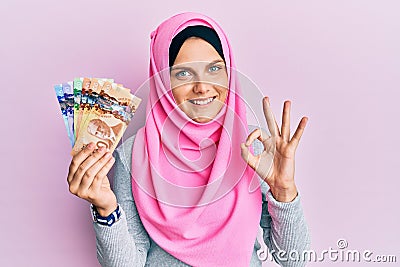 Young caucasian woman wearing islamic hijab holding canadian dollars banknotes doing ok sign with fingers, smiling friendly Stock Photo