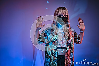 Young adult woman posing in a bright neon studio wearing protective medical mask and looking frightened holding hands up Stock Photo