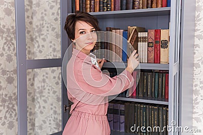 Young caucasian young woman with brunette hair taking boor for reading on free time standing near bookcase, positive Stock Photo