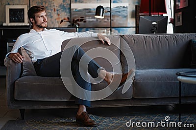 Young and charming businessman is thinking about business while sitting on the sofa in his modern office. Stock Photo