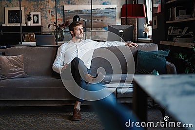 Young and charming businessman is thinking about business while sitting on the sofa in his modern office. Stock Photo