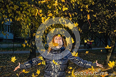Young cheerful cute girl woman playing with fallen autumn yellow leaves in the park near the tree, laughing and smiling Stock Photo