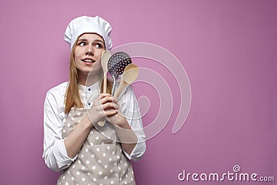 Young cheerful girl cook in apron holds spoons and kitchen items and dreams on a pink background, portrait of a housewife woman Stock Photo