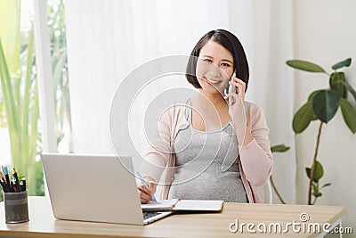 Young cheerful pregnant woman talk on phone. Also working remote using laptop Stock Photo