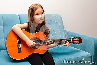 Young child, little school age girl playing classic acoustic guitar, practicing musical instrument sitting on the sofa Stock Photo