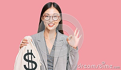 Young chinese woman wearing business suit holding dollars bag doing ok sign with fingers, smiling friendly gesturing excellent Editorial Stock Photo