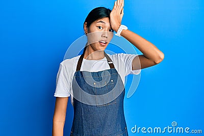 Young chinese woman wearing waiter apron surprised with hand on head for mistake, remember error Stock Photo