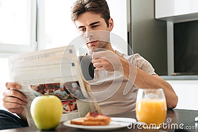 Young concentrated man reading newspaper while sitting in kitchen Stock Photo