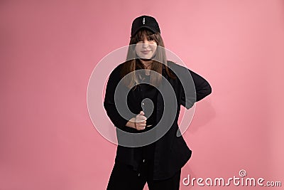 Young, confident woman dancer in black baseball cap, shirt and sportswear on pink background in studio looks confidently into Stock Photo