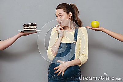 Young confused pregnant woman choosing between apple and cream cake Stock Photo