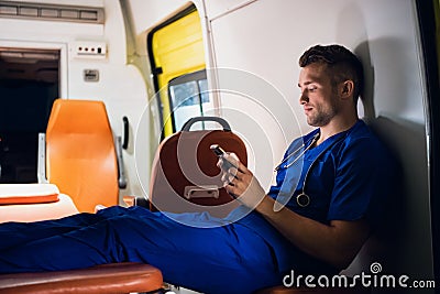 Young corpsman in blue medical uniform sits with phone in his hands in the ambulance car Stock Photo