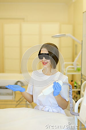 Young cosmetologist wearing special purpose glasses and latex gloves sitting near permanent makeup device. Stock Photo