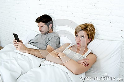 young couple in bed unsatisfied wife bored frustrated and angry while internet addict husband is using mobile phone social networ Stock Photo