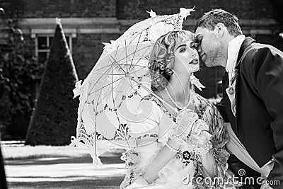 Young couple dressed in vintage costume relaxing with man on one knee, kissing on lawn in front of stately home Stock Photo