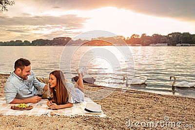 Young couple enjoying a picnic at the beach. Lying on the picnic blanket. White swans swimming the background Stock Photo