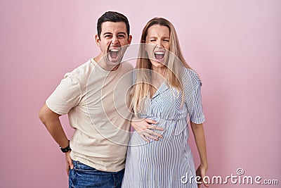 Young couple expecting a baby standing over pink background angry and mad screaming frustrated and furious, shouting with anger Stock Photo