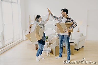 Young couple five high five to each other, carry big cardboard boxes during moving day, agree to work as team, pose in new Stock Photo
