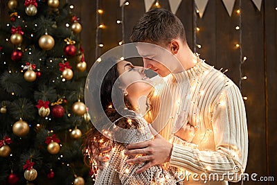 Young couple kissing in christmas lights and decoration, dressed in white, fir tree on dark wooden background, romantic evening, w Stock Photo