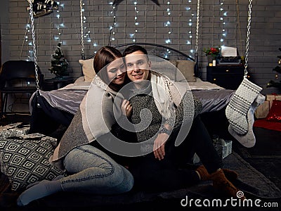 Young couple in love,sitting near bed with christmas lights behind, embracing, kissing. Brunette man and woman, wearing grey Stock Photo