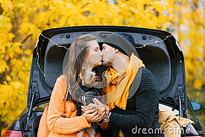 A young couple in love is sitting on the open trunk of a black car with their little dog in the autumn forest. Lovers kiss, and Stock Photo