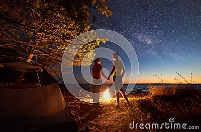 Young couple man and woman having rest at tourist tent and burning campfire on sea shore near forest Stock Photo