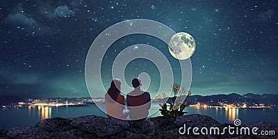 young couple man and woman sit on rock at night sea watching starry sky and big moon on horizon mediterranean city Stock Photo