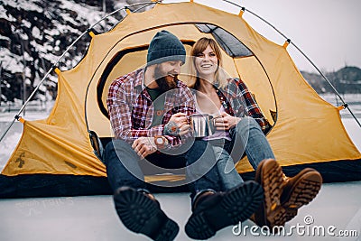 Young couple sits in tent, smiles and drinks hot tea during winter hike. Stock Photo