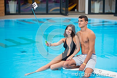 Young couple sitting on the edge of the swimming pool and taking selfie photo on the phone with selfie stick Stock Photo