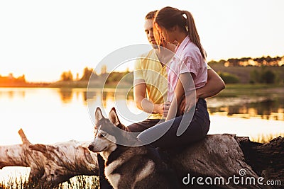 A young couple is sitting by the lake at sunset with a Husky breed dog. Stock Photo