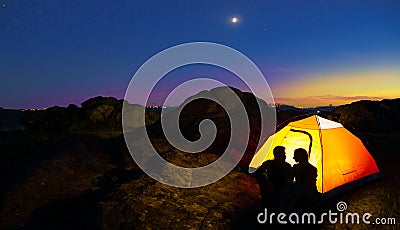 Young Couple Sitting near Illuminated Tent and Looking at Each Other at Beautirul Evening in the Mountains Stock Photo