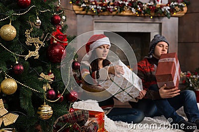 Young couple sitting on a rug in front of the decorated fireplace and Christmas tree unwrapping their Christmas gifts Stock Photo