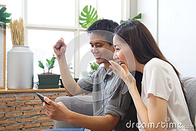 Young couple sitting on sofa are watching mobile phone and feeling surprise&happy. Stock Photo