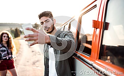 A young couple with smartphone by a car on a roadtrip through countryside, taking selfie. Stock Photo