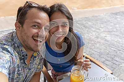 Young couple taking a selfie with smart phone while drink a smoothy and a coffee. Husband and Wife laughing, sitting in a bar. Stock Photo