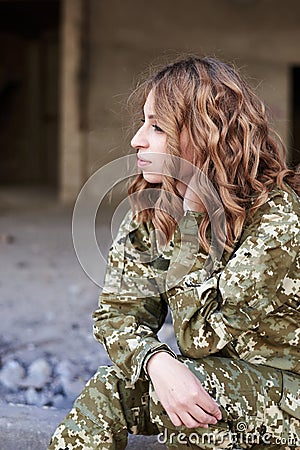 Young curly blond military woman, wearing ukrainian military uniform, sitting on pavement edge. Close-up portrait of army female Stock Photo
