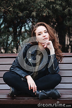 young curly stylish woman wearing black jacket sitting on a bench in city Stock Photo