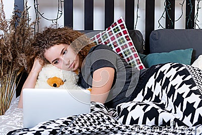 A young curly teen lies in bed caught a teddy bear and looks at the laptop screen Stock Photo