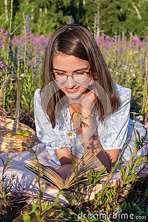 A young cute attentive girl lies on the green grass and reads a book illuminated by the setting sun. Stock Photo