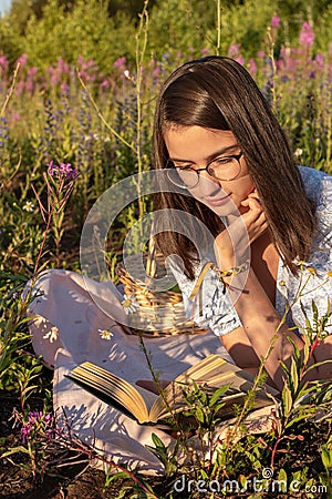 A young cute attentive girl lies on the green grass and reads a book illuminated by the setting sun. Stock Photo