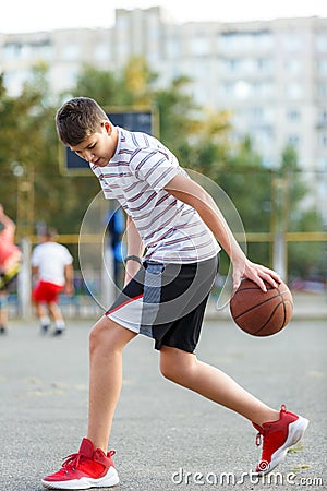 A young cute boy plays basketball at the outdoor streetball court on a sunny summer day. Teenager player in action dribbling Stock Photo