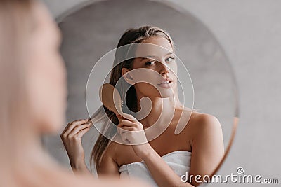 Young cute lady wrapped in towel, standing in front of mirror at bathroom and combing her hair after shower Stock Photo