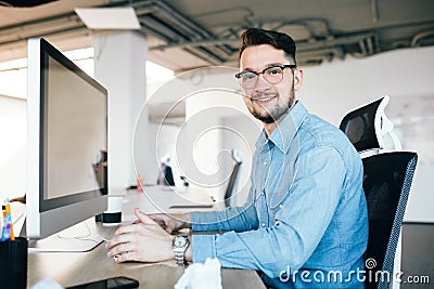 Young dark-haired man in glasses is working with a computer at his desktop in office. He wears blue shirt and smiles to Stock Photo