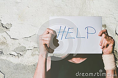Young depressed homeless man with bandage on his hand from suicide attempt holding help sign written on paper while he leaning his Stock Photo